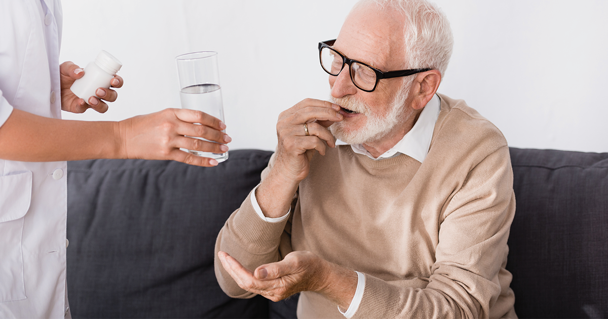 Elderly man putting medication in his mouth while health care practioner hands him a glass of water.