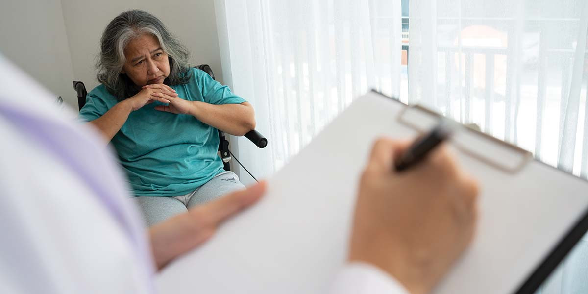 closeup of hand with pen and clipboard with elderly patient sitting in the background