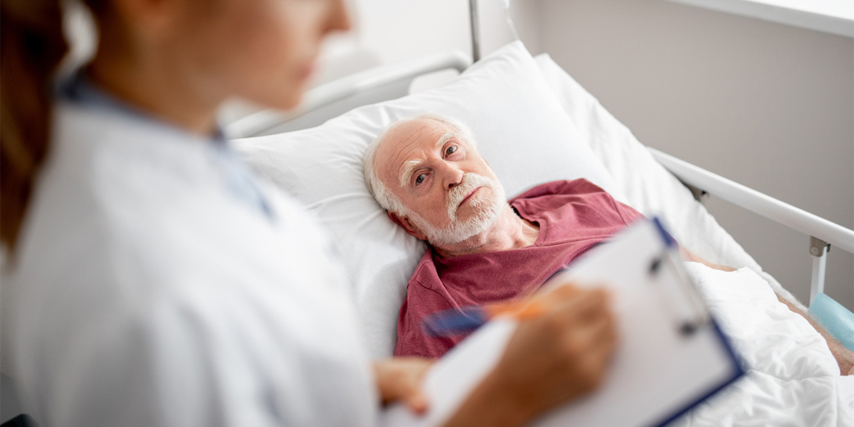 elderly man lying in hospital bed with healthcare working holding a clipboard