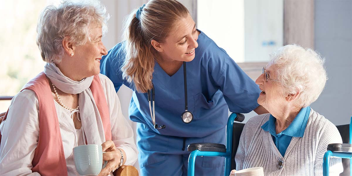 two elderly women enjoying coffee with healthcare worker checking on them.