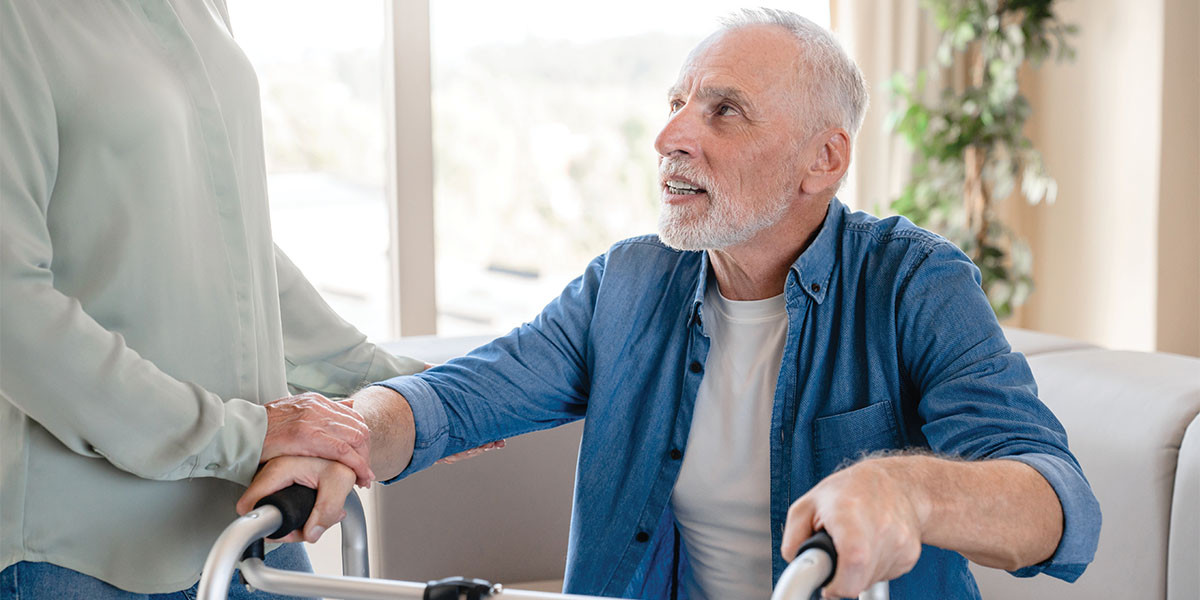 older adult man seated and looking up to adult, with hands on walker, getting ready to get up from seat