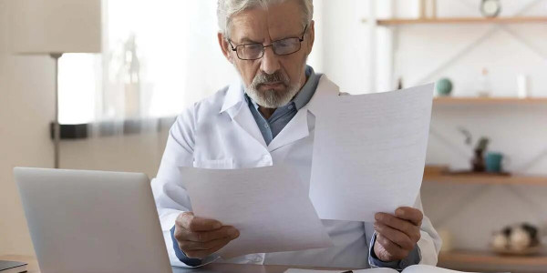 Healthcare worker with a white coat holding papers seated in front of laptop computer.