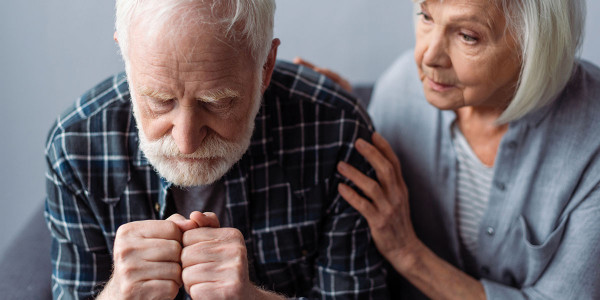 Elderly gentleman sitting with hands together in front of him with concerned elderly woman sitting next to him, touching his shoulder and arm