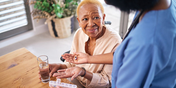 older adult woman with a glass of water and weekly medication box dispenser being handed medications by healthcare worker