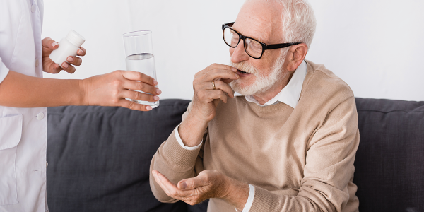 Elderly man putting medication in his mouth while health care practioner hands him a glass of water.