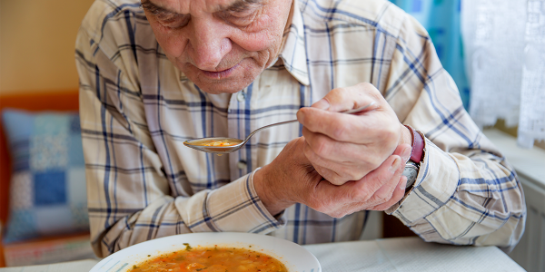 elderly man eating soup, holding left hand with right to help steady tremors