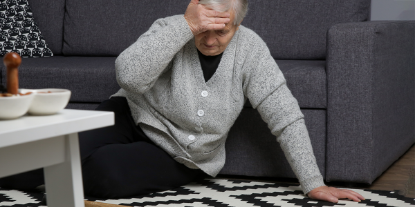 Elderly woman on floor in front of couch with one hand supporting her on floor and one hand on her head.